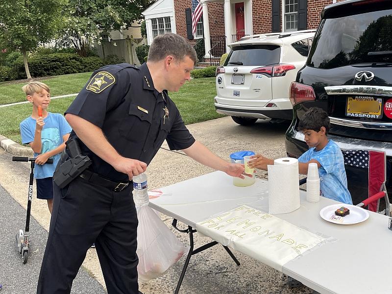 sheriff reaching for a cup at a table with a little boy and a sign that says "Free Lemonade"