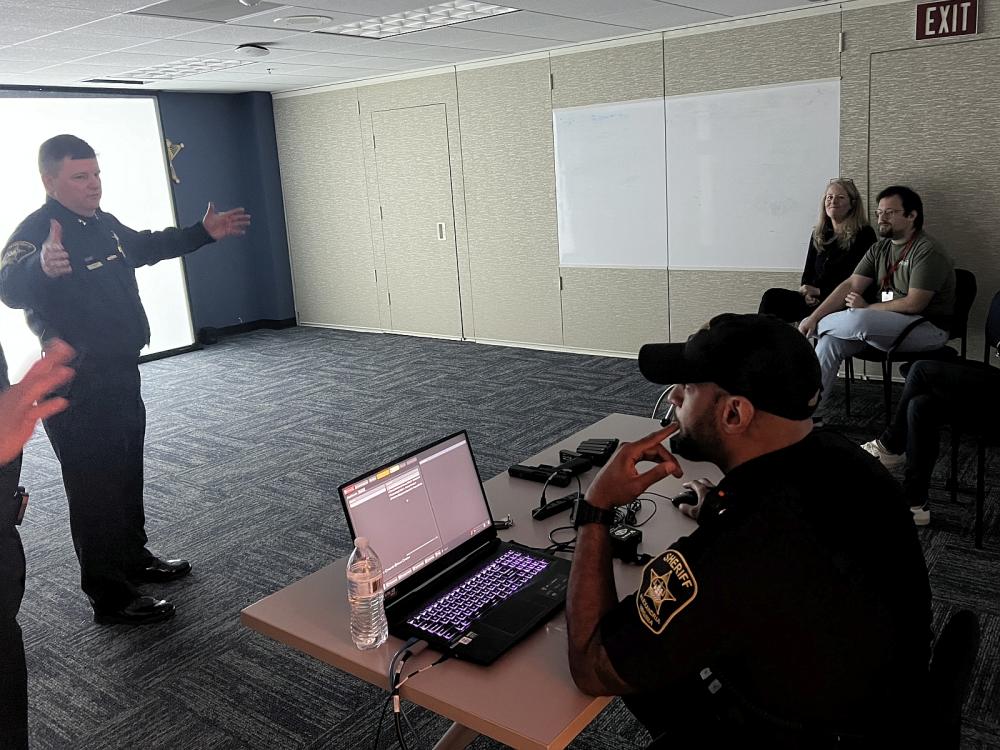 Uniformed Sheriff's Office commander speaking to three civilians in a dark room with a white screen. Another deputy operates at laptop.