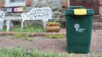 Photo of a  green residential curbside compost collection can in front of a white metal bench