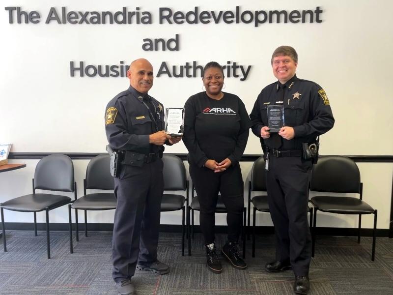 Uniformed deputy on left holding glass plaque, civilian wearing shirt that says ARHA and Sheriff in uniform also holding a glass plaque standing in front of lettering on the wall that reads The Alexandria Development and Housing Authority