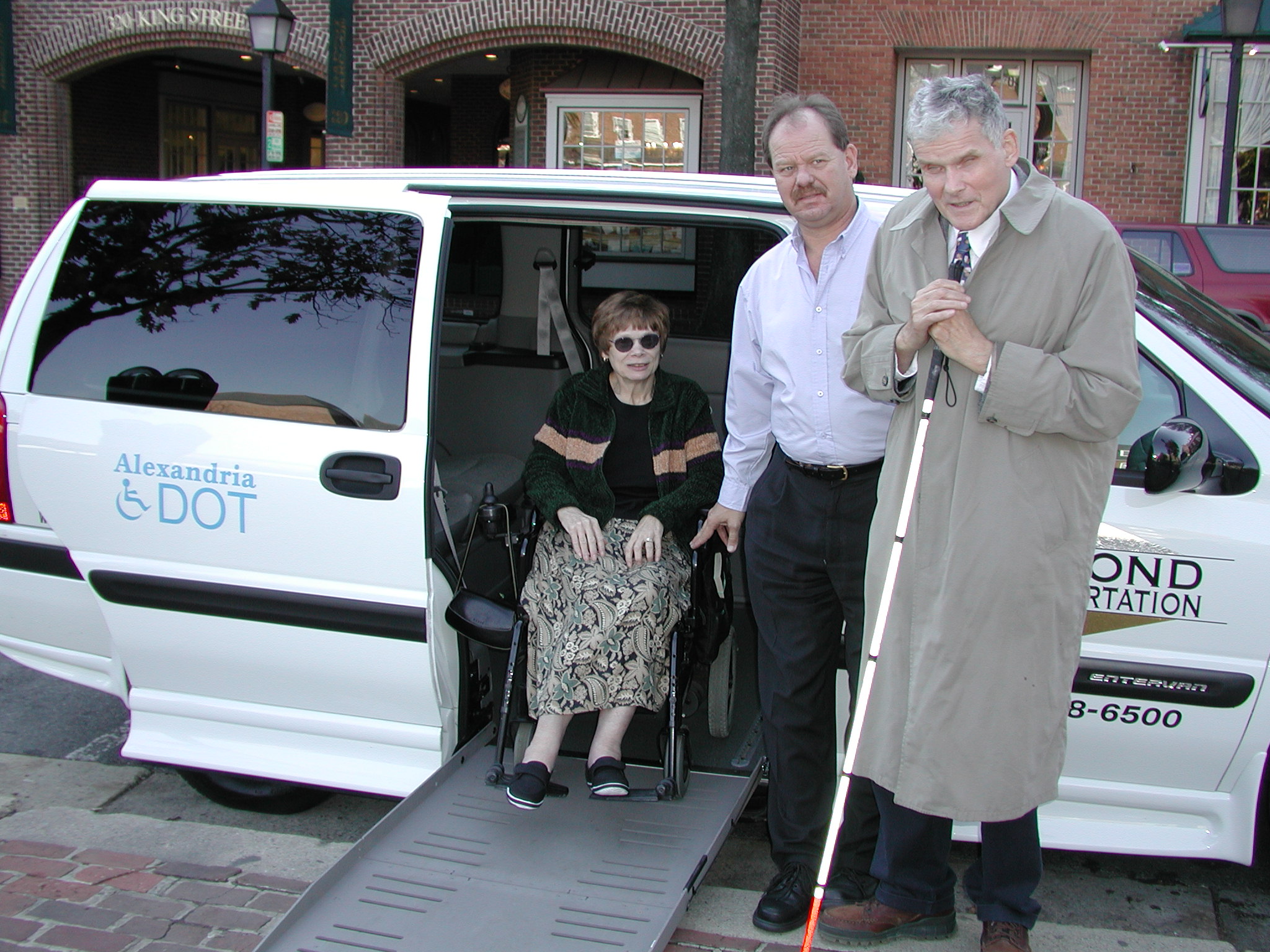 Photo of two DOT Paratransit users (woman in wheelchair, man with white cane) and their driver (man in white shirt) in front of a white DOT van. 