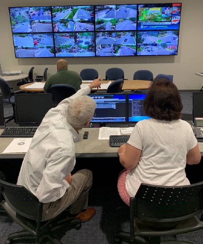 Two traffic center employees with their backs to the camera points at the eight-screen display that shows traffic on streets around Alexandria