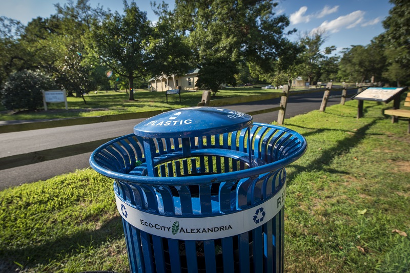 a photo of a blue, City-issues metal recycling can in an Alexandria park. "Eco-City Alexandria" sticker affixed to the front. 