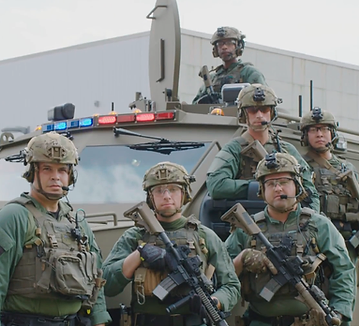 Six men in combat gear and holding weapons pose in front of a military vehicle.