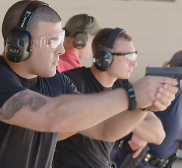 A man in the foreground, wearing eye and ear protection, aims a handgun 