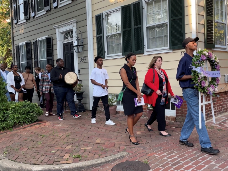 a man carrying a memorial wreath and leading a procession of city officials and community members along a brick sidewalk