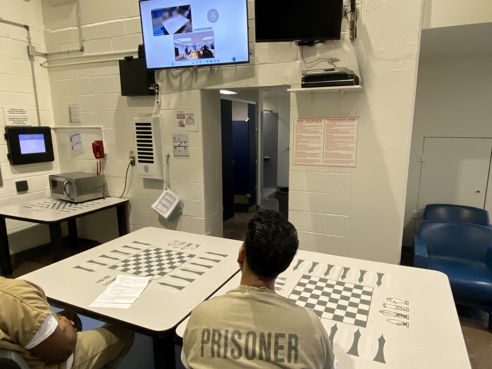 Two inmates, visible from the back, wearing tan jumpsuits that say Prisoner, seated and interacting with students on a television screen on a wall