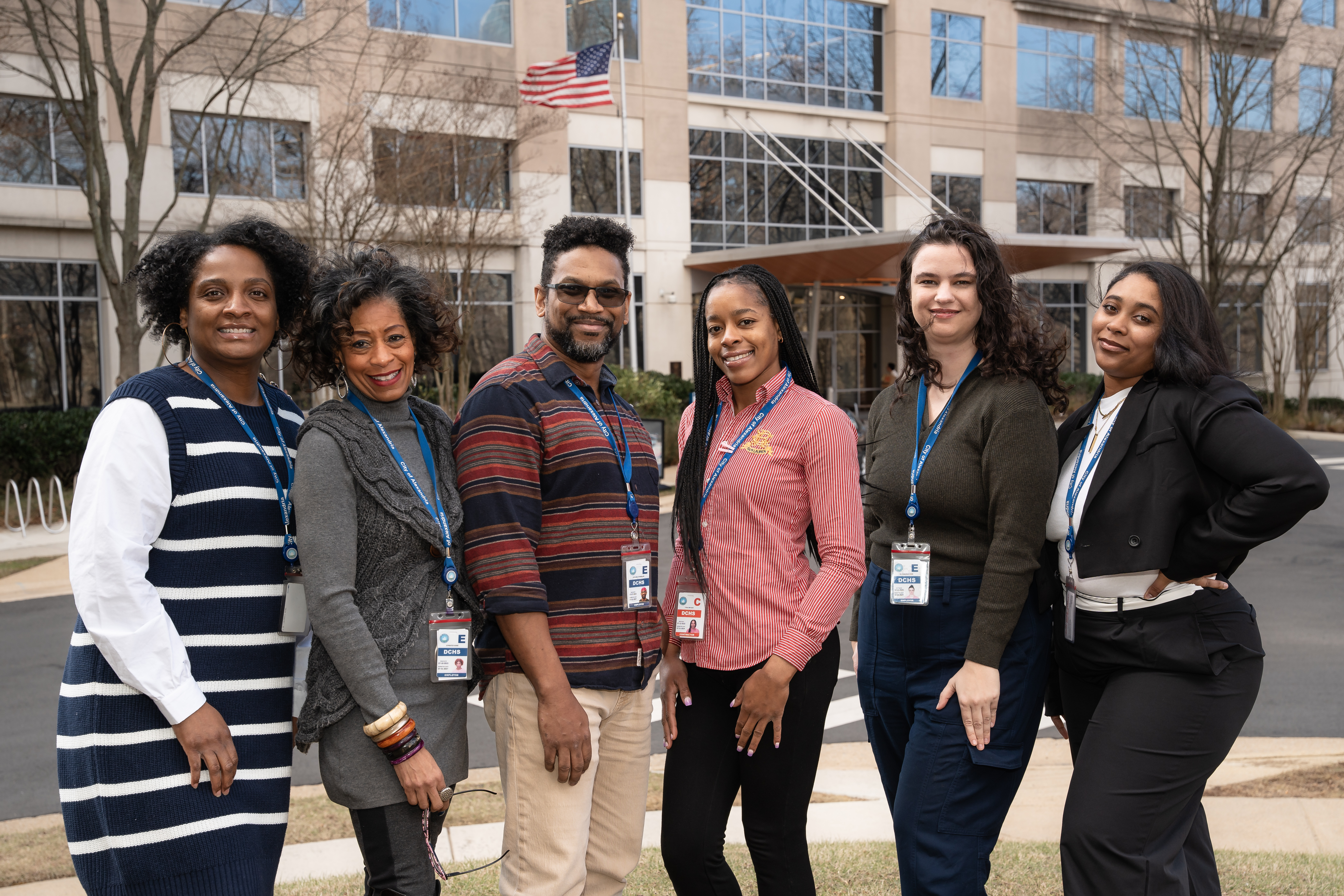  A group of six people is standing outdoors in front of a modern office building. They are all wearing lanyards with ID badges.