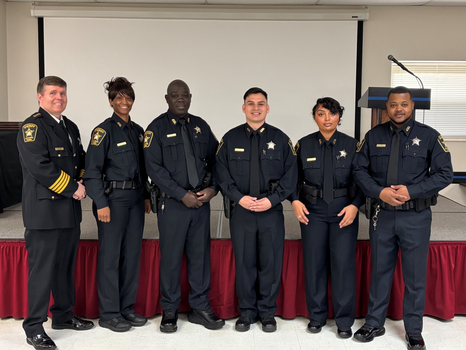 Sheriff in formal uniform standing next to five recruit deputies in blue uniforms at their graduation