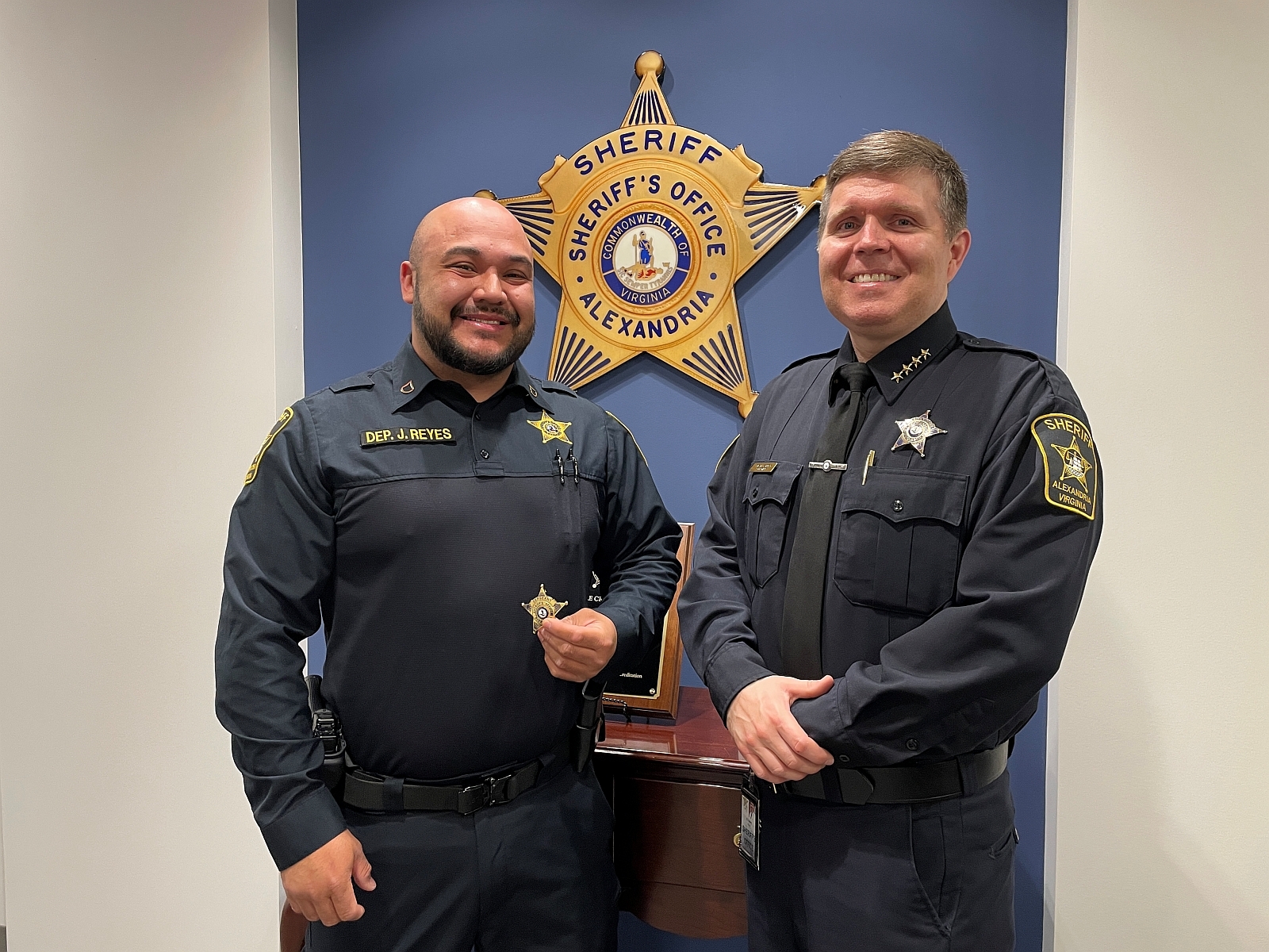 Deputy and Sheriff, both in blue uniforms, standing in front of a wall with a large gold sheriff's star