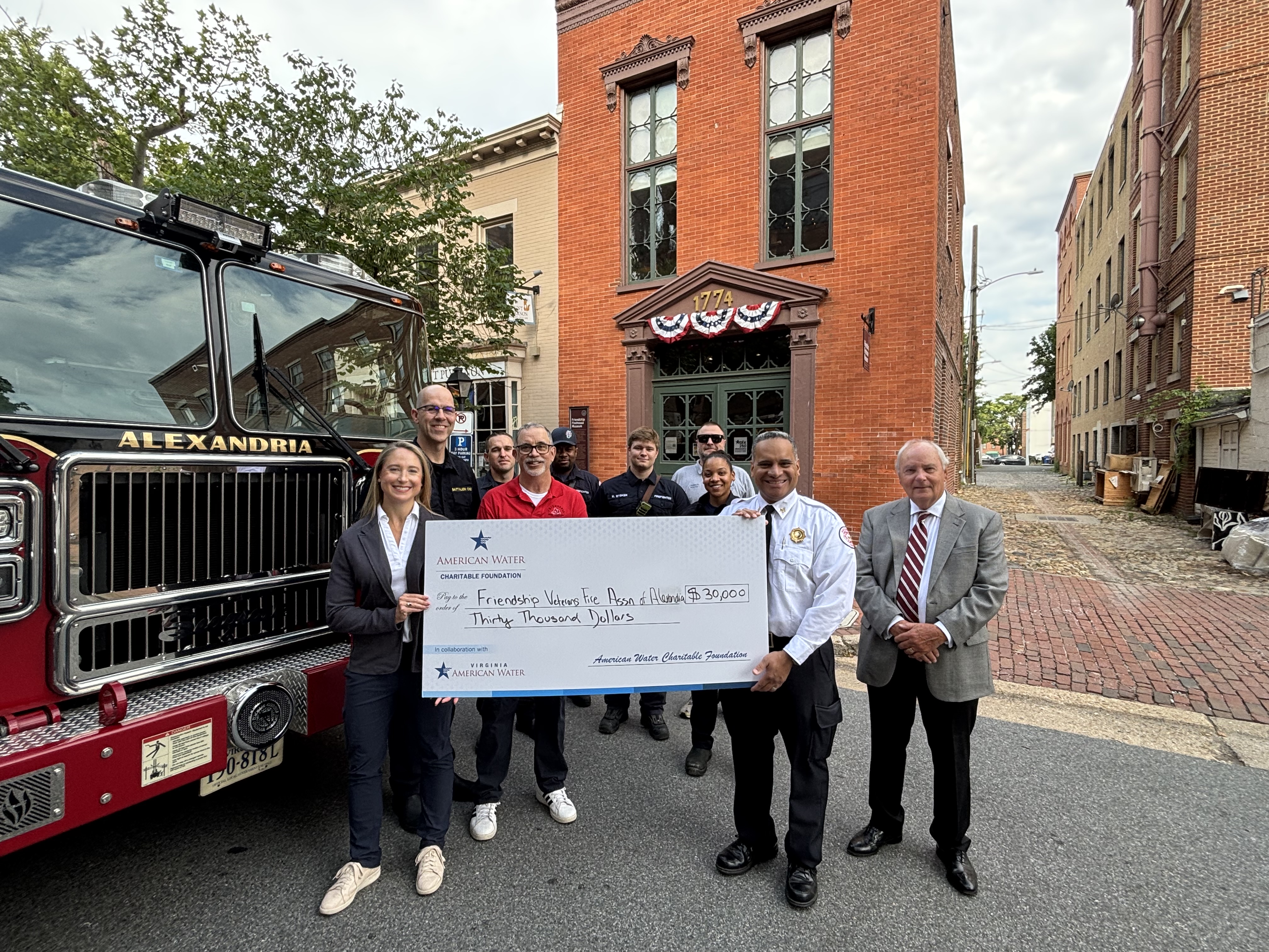 Group of 10 people smiling holding a large check from American Water. The amount is for thirty thousand dollars. You can partially see a fire engine on the left and brick building in the back and an alley way. The group is standing on the street.