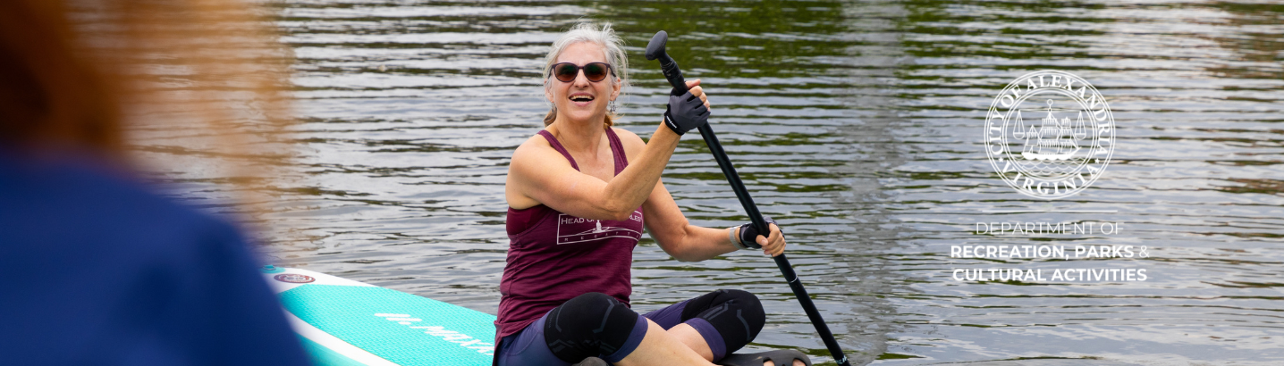 Woman in a maroon tank top, wearing black sunglasses, sitting on a blue paddleboard while holding a paddle and smiling wide towards a person who's blurred on the left side of the image