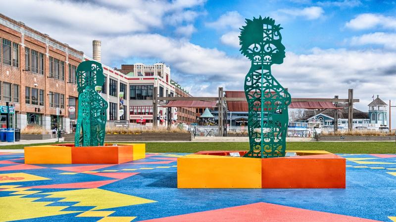 Two of four figures in Wrought, Knit, Labors, Legacies. Metal figures painted green and adorned with scroll cuts throughout. The ground mural is blue, yellow, and orange.
