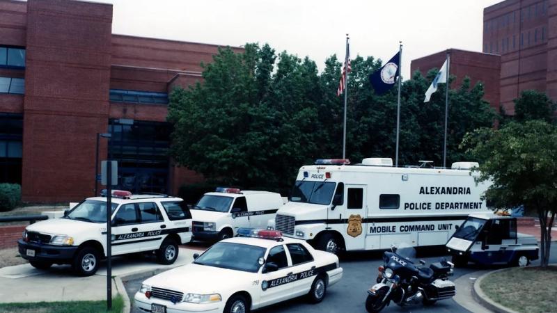 A color photo of a parking lot with a collection of APD vehicles (from the top, clockwise): mobile command unit, small utility vehicle, sedan, SUV, box van in front of a dark red brick building