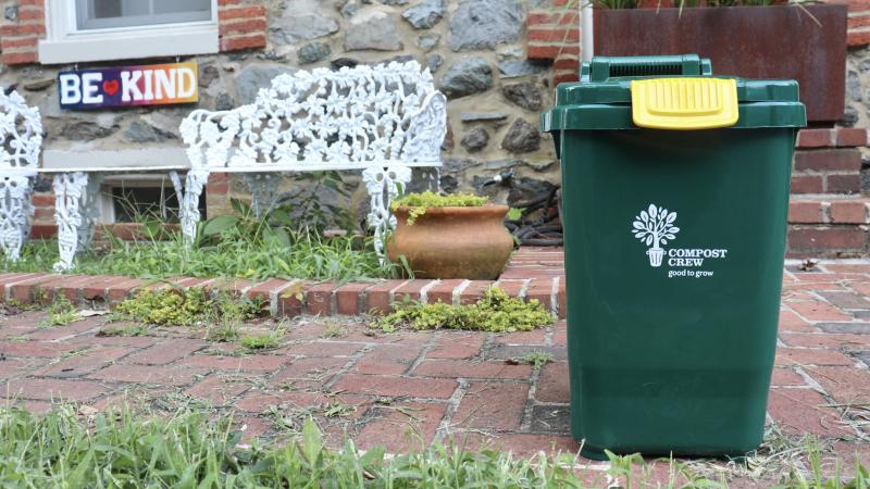 Photo of a  green residential curbside compost collection can in front of a white metal bench