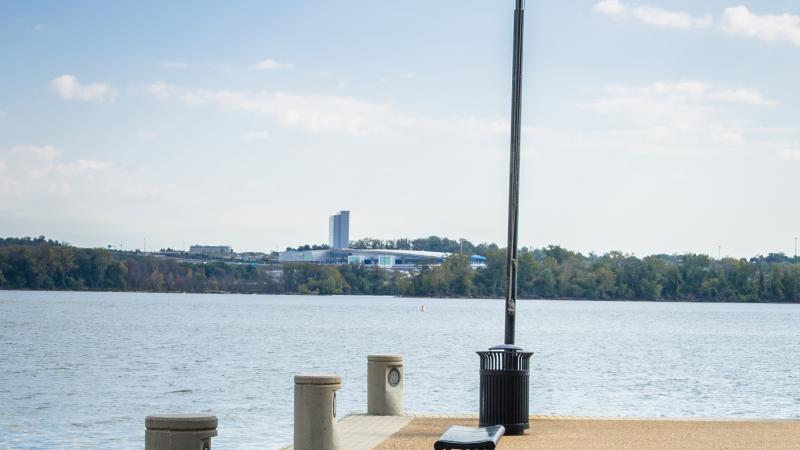 The end of a pier at the Waterfront juts out into the Potomac. A short bench is in the foreground and an American flag waves on a flagpole at the end of the pier.