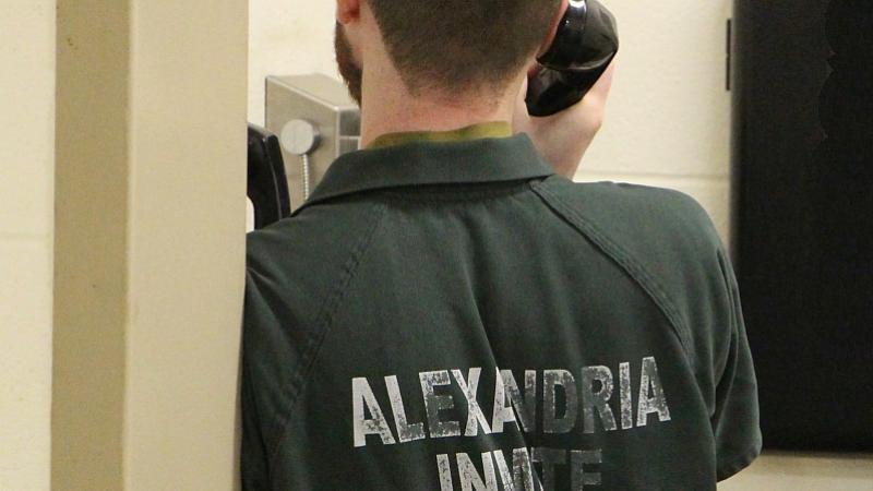 view from behind of an inmate wearing a green jumpsuit that says "Alexandria Inmate" and holding a phone to his ear