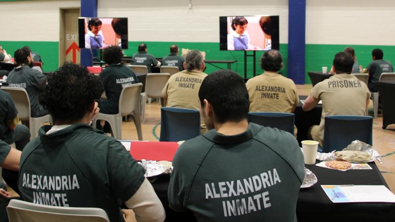 Visible from the back, several inmates wearing tan or green jumpsuits and seated at tables eating lunch and watching a movie on two screens.