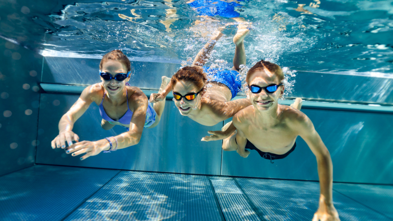 Three teens swimming underwater in an indoor pool with goggles on and smiling