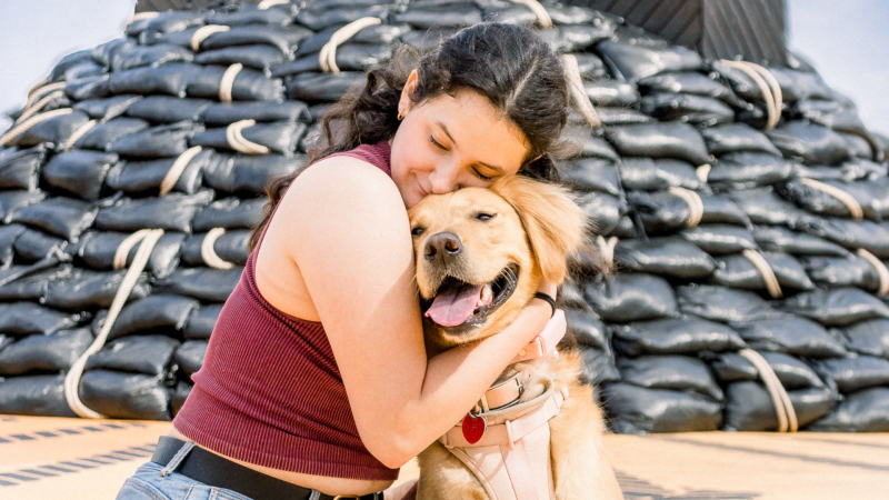 Image of a young lady in maroon top smiling with her eyes closed while hugging her dog with golden fur posing in front of the Break Water installation