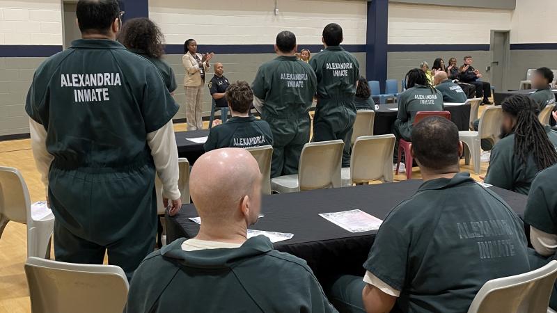 Several inmates in green jumpsuits, some standing and seated, in a gymnasium visible from behind