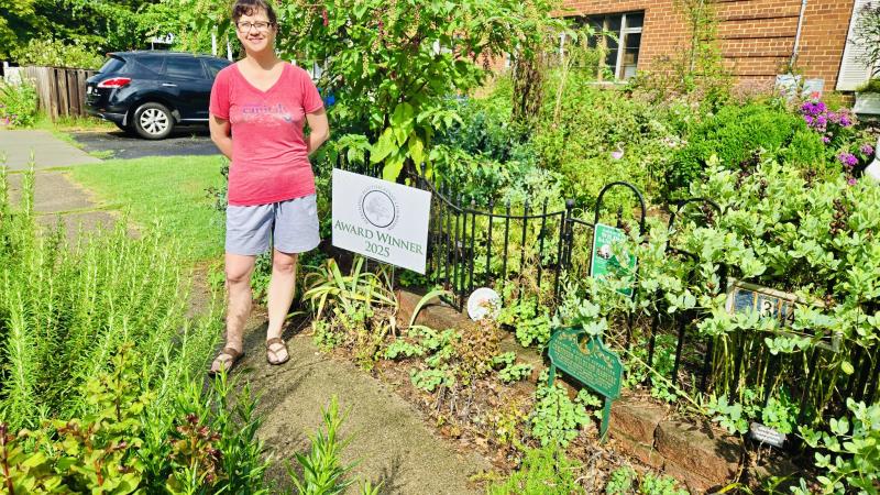 Image of a the Award Winner in front of the sign and surrounded by green shrubbery and native plantings