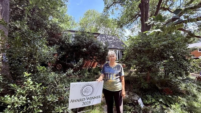 Woman standing next to a yard sign that says Award Winner 2025 in front of a very green yard