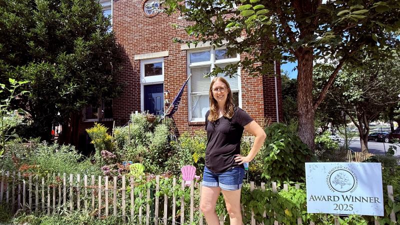 A woman stands in front of her fence surrounding a corner brick townhouse with a blue door and tall shrubs and trees and lots of native plantings
