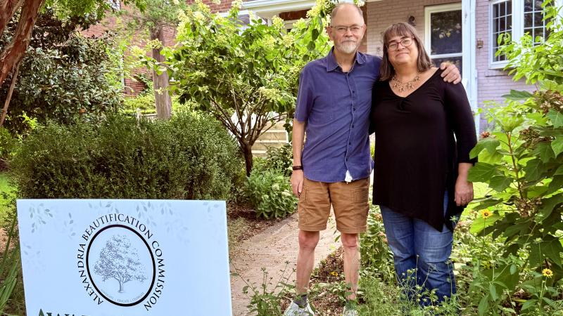A man and a woman stand behind a sign that says Award Winner 2025 located in their front yard that's very green and shady
