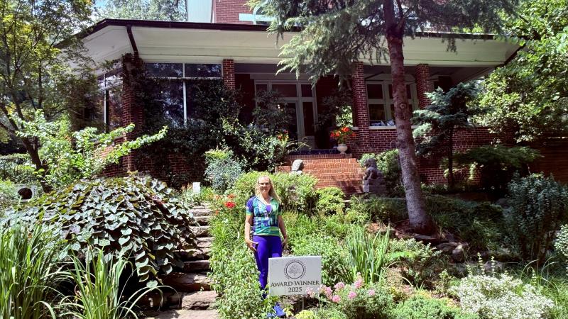 Woman stands behind a sign that says Award Winner 2025 posing in front of her house in the front yard full of native plantings and greenery