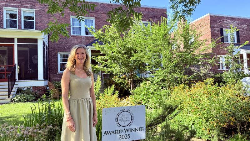 Winner of the Beautification Street Award stands in front of her front yard full of green trees and native plantings