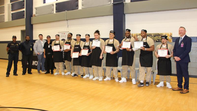 14 people standing next to each, includ a deputy, two kitchen managers, two people in business attire, and nine inmate workers wearing tan uniforms, black aprons and chef's beanies and holding certificates