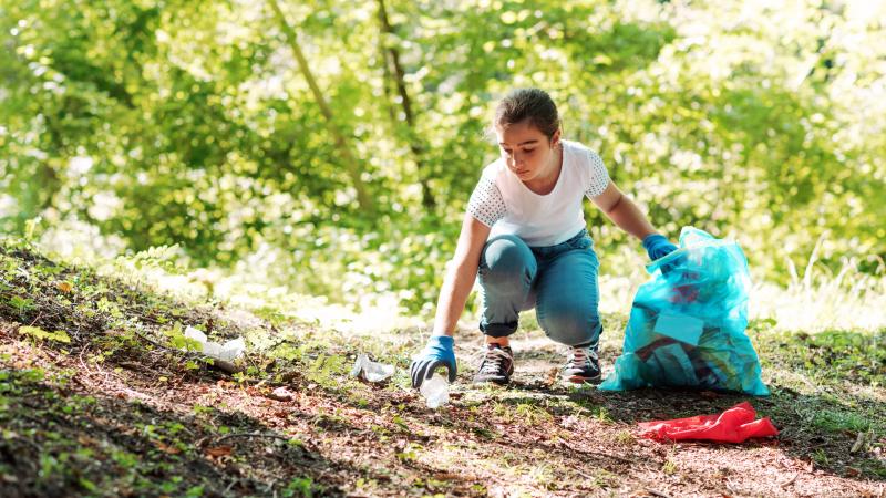 Girl dressed in white shirt and jeans picking up trash in the woods while holding a blue trash bag