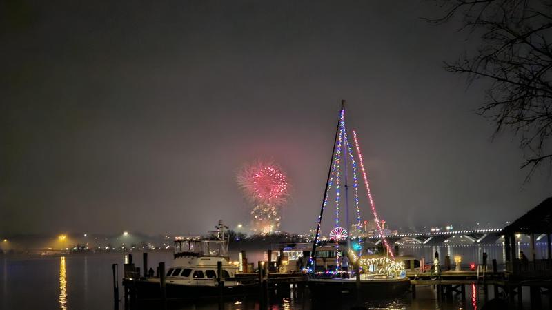 Fireworks in the distance behind a sailboat with holiday lights strung along its rigging.