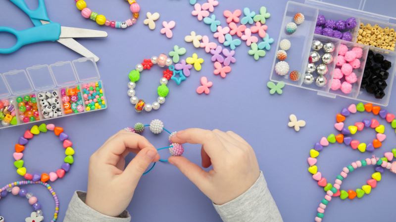 A pair of hands engaged in making a bracelet with bracelet-making materials including colorful beads and scissors are scattered on a purple surface