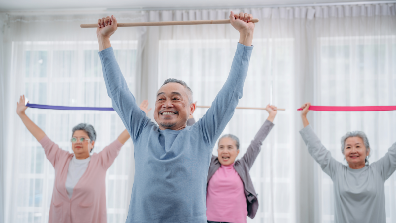 Image of four seniors smiling while they have their hands straight up in the air and holding thing wooden dowels and rubber exercise bands