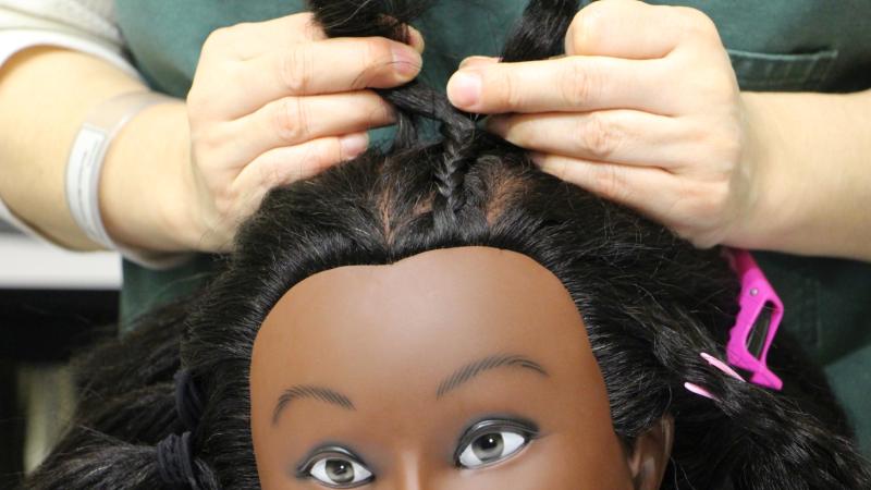 Close up of a mannequin head with a student's hands braiding cornrows