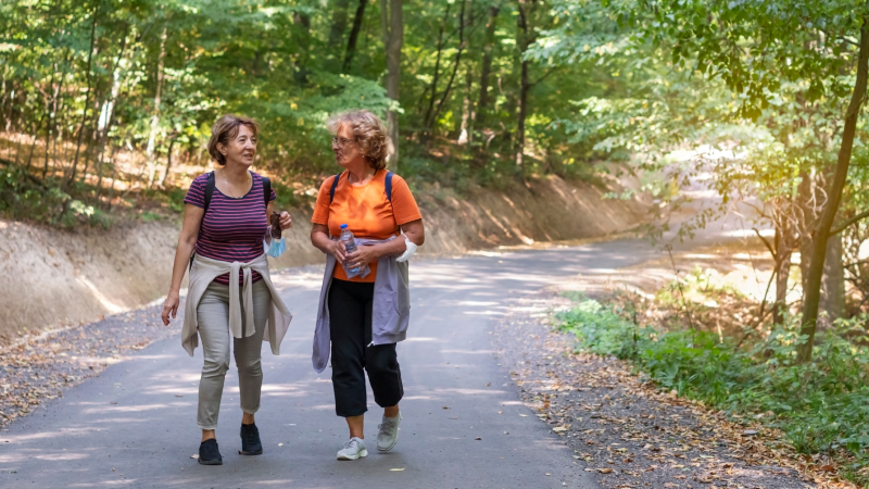 Two women with sweaters wrapped around their waists engaged in conversation while walking on a paved path surrounded by trees and shrubs