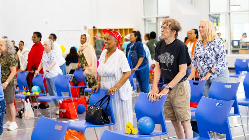 A group of seniors exercising using chairs and exercise balls. They're all standing inside the Patrick Henry gym
