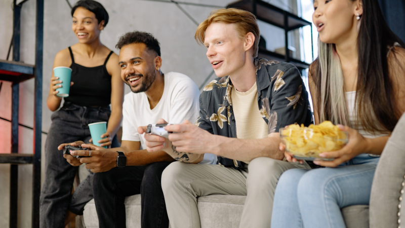 Two male adults engaged in a video game and two female adults watching while holding cups and chips