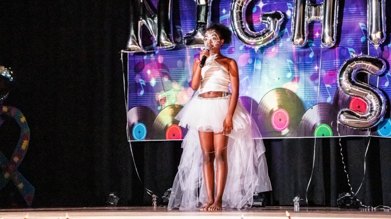 Performer in a white tulle dress with a white painted face and holding the mic