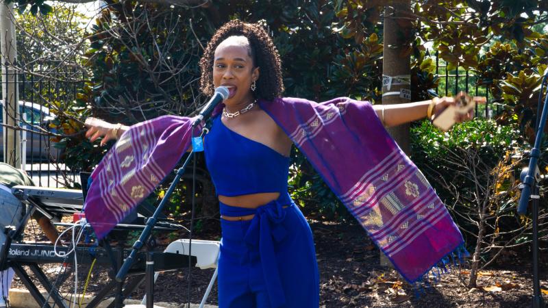 woman in blue jumpsuit with purple decorative shawl arms outstretched in front of a microphone