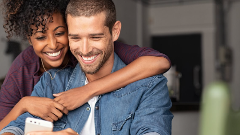 Smiling couple looks at a smartphone together. Text reads: CommonWealthONE. Live Richer. Even on the same paycheck!.