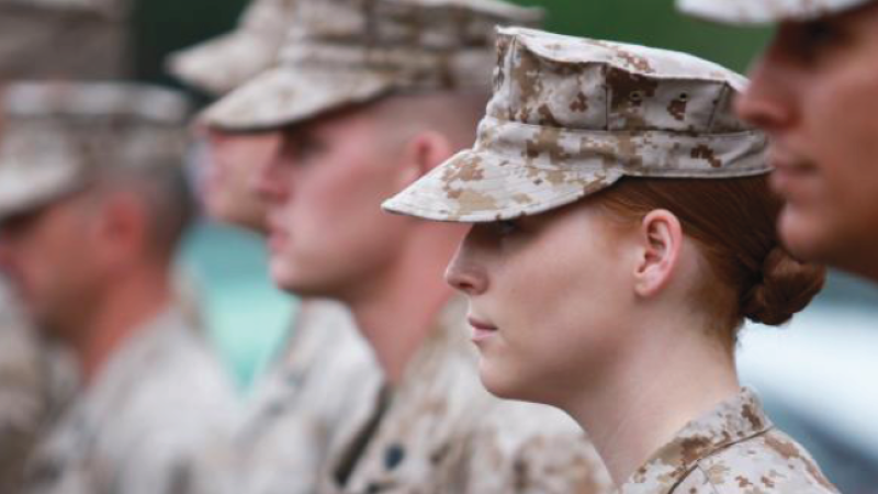 A group of soldiers in camouflage uniforms and caps stand in a line, facing forward, with one woman in focus among them.