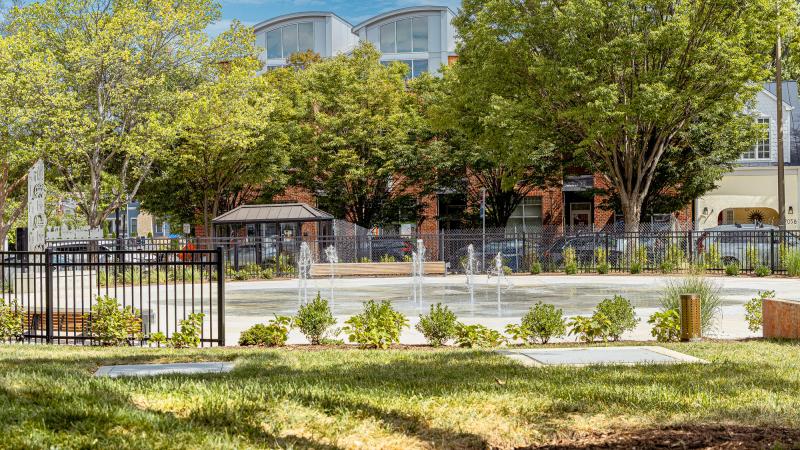 Colasanto spray park with the fountain on, green trees and bushes surrounding the fountain along with seating and black fence