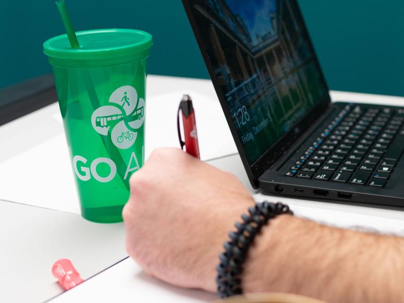 A photo of a person's desk (a hand writing, a laptop, and a GO Alex cup)