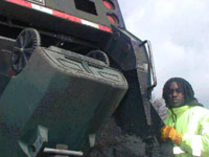 Photo of person standing beside a trash truck as a residential trash cart is emptied