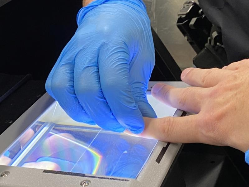 close up of hands wearing blue gloves holding another hand on a glass scanner to record fingerprints