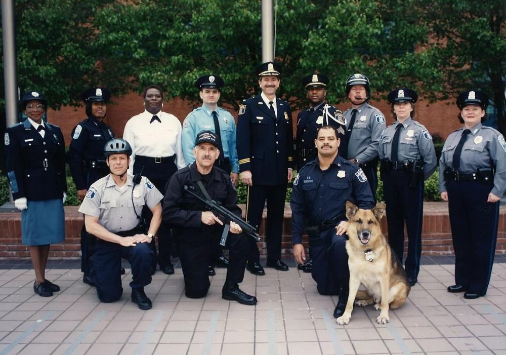 A color photo of a group of Alexandria police officers and a K9 officer on a stone patio in front of a pair of flagpoles. The group is comprised of officers representing a variety of roles in the Police Department.
