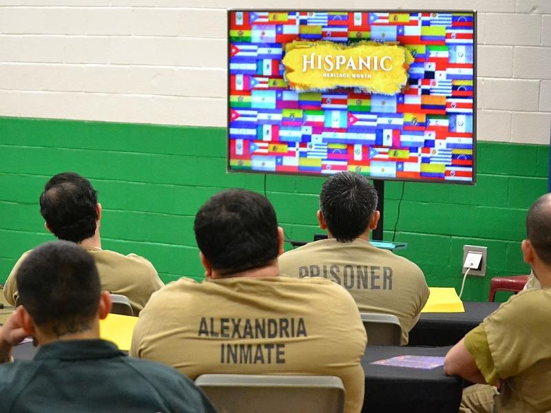 backs of five inmates, seated and wearing tan or green jumpsuits, looking at a large televsion screen with colorful flags displayed